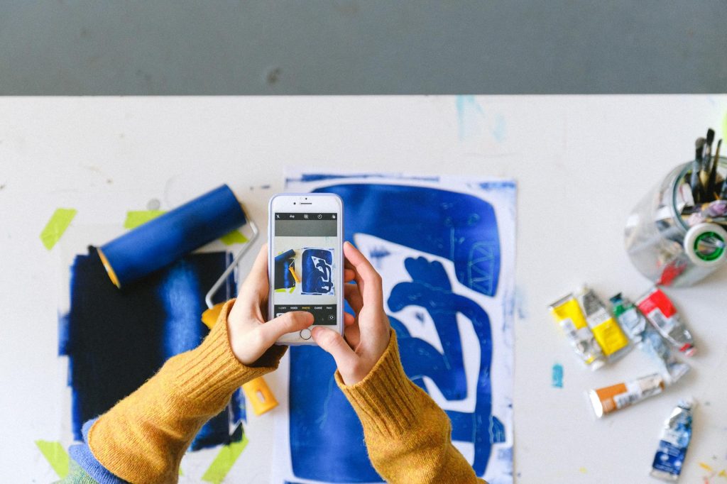 Artistic workspace with hands capturing artwork photo, surrounded by paint and tools.