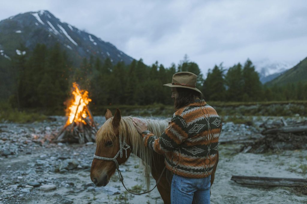 A man in a patterned jacket and hat pets a horse near a campfire set against a mountainous backdrop.