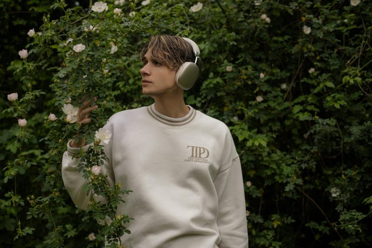 A young man wearing headphones and a white sweatshirt stands amidst lush greenery and flowers.
