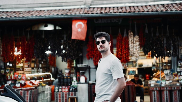 Man wearing sunglasses, standing in front of a vibrant traditional Turkish market.