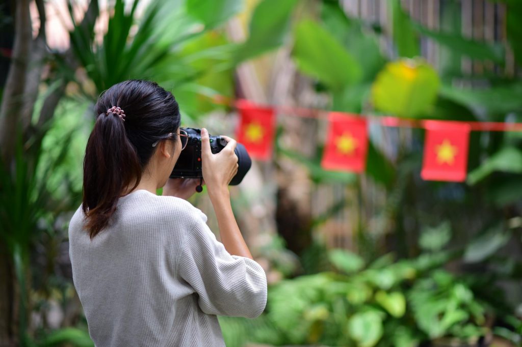 A woman takes photos in a lush outdoor setting with flags in the background.