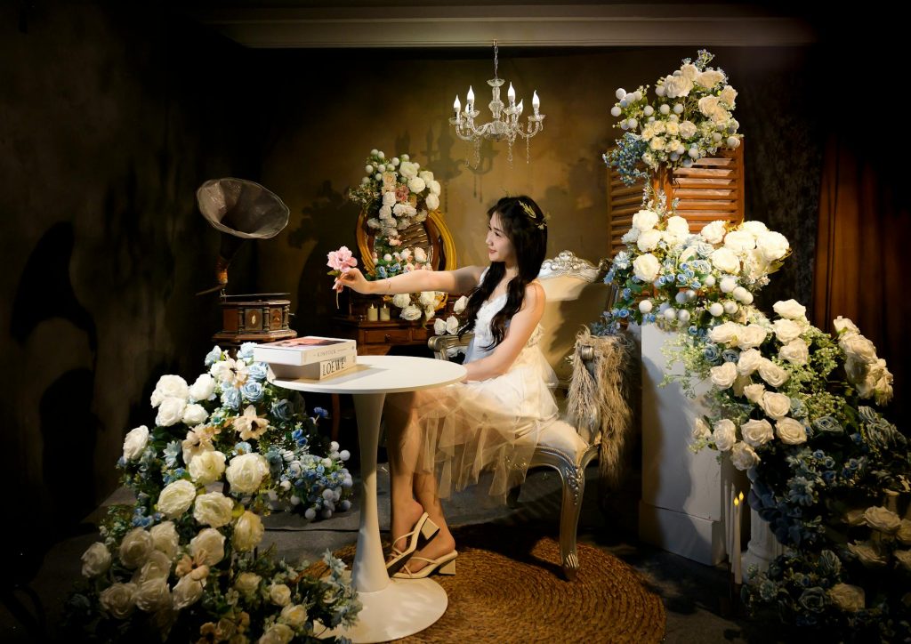 A woman in a floral setting holds a pink flower, surrounded by elegant decor and soft lighting.