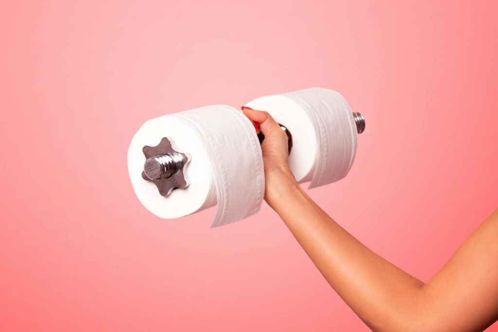 A playful image showing a hand holding a dumbbell made of toilet paper against a pink background.