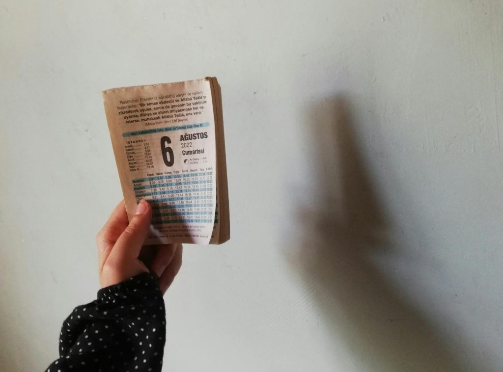 A hand holds a calendar page showing August 6, 2022, casting shadow on wall.