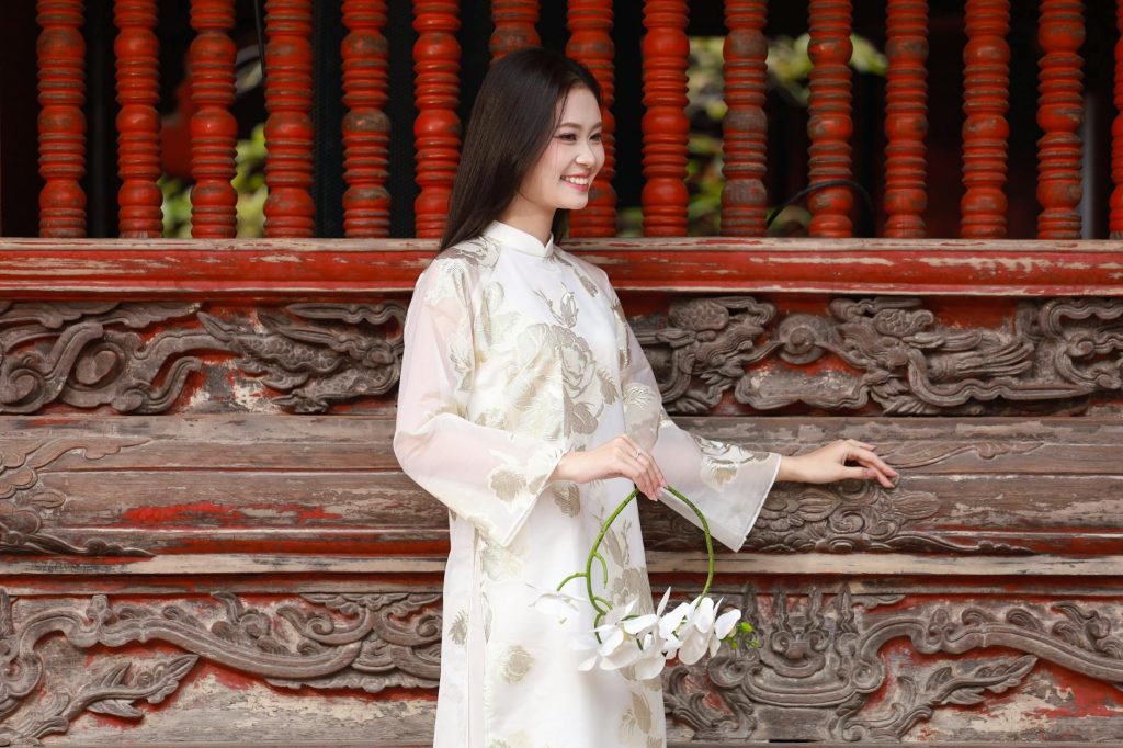 Young woman in traditional Vietnamese áo dài holding lotus flowers, smiling against ornate wooden backdrop.