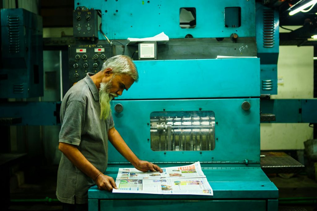 An older man examines freshly printed materials at a large, teal printing press.