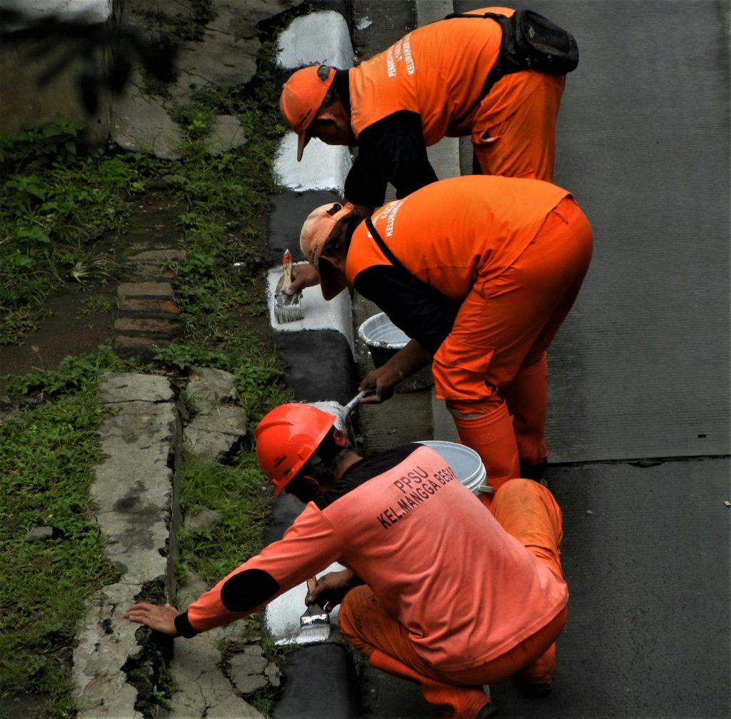Three workers painting a curb in Jakarta, wearing orange uniforms, in a street maintenance task.