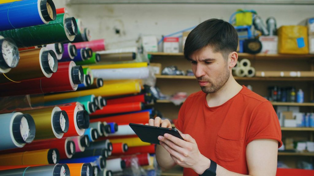Man using a digital tablet in a print shop with colorful rolls of paper.