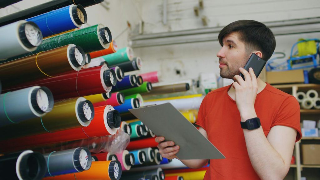 Young man talking on phone, holding a clipboard in a print shop filled with colorful rolls.