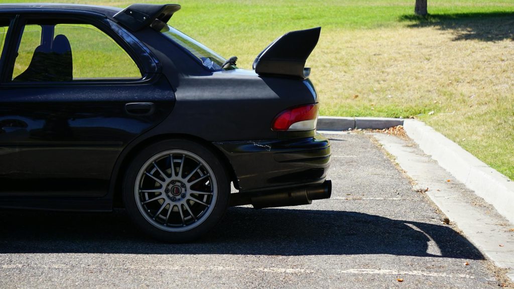 Black Subaru Impreza with spoiler parked outdoors, sunlight casting shadows.